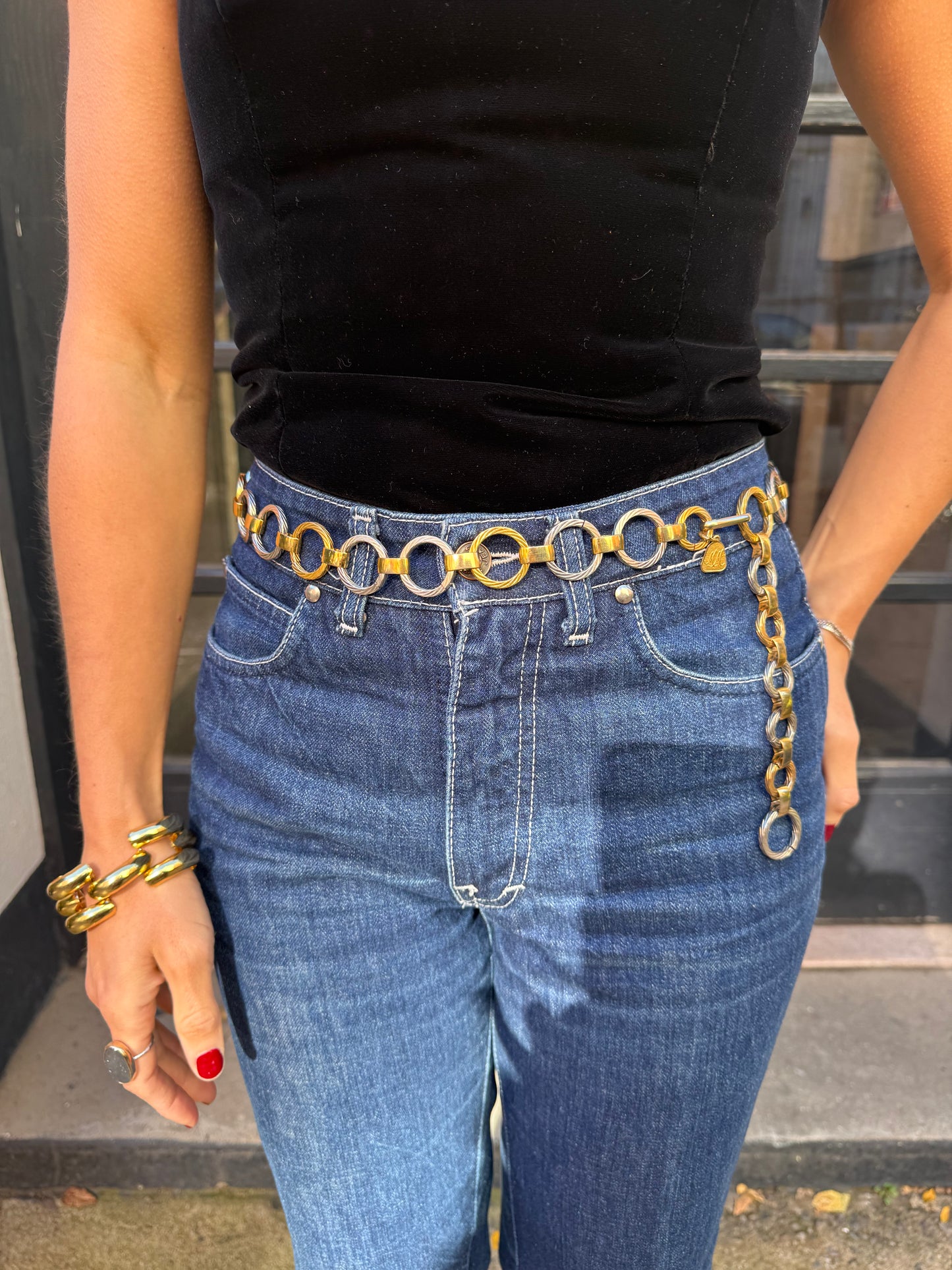 Person wearing blue jeans with gold chain detail and a black top, standing indoors.